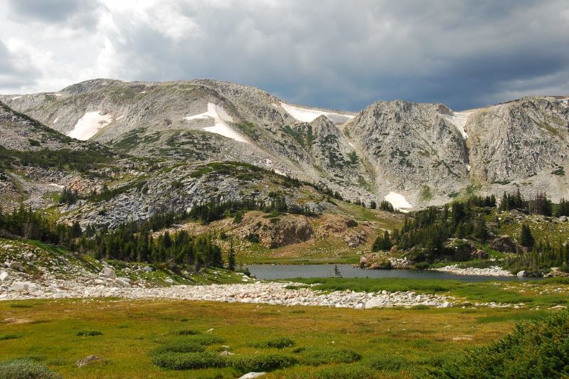 A small lake below snowy mountains, sparse forests and rocks.